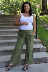 woman wearing a white tank top and olive green maternity wide-leg pants, holding her baby bump outdoors