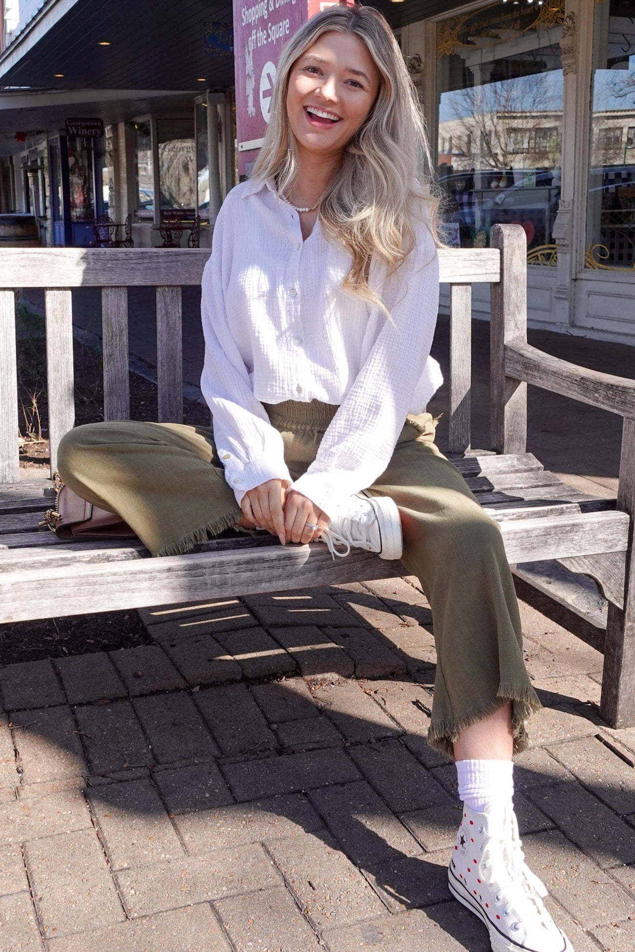 woman sitting on a bench wearing a white long sleeve button-up shirt and olive green culottes, with white sneakers and a smile
