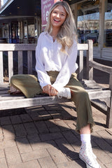 woman sitting on a bench wearing a white long sleeve button-up shirt and olive green culottes, with white sneakers and a smile