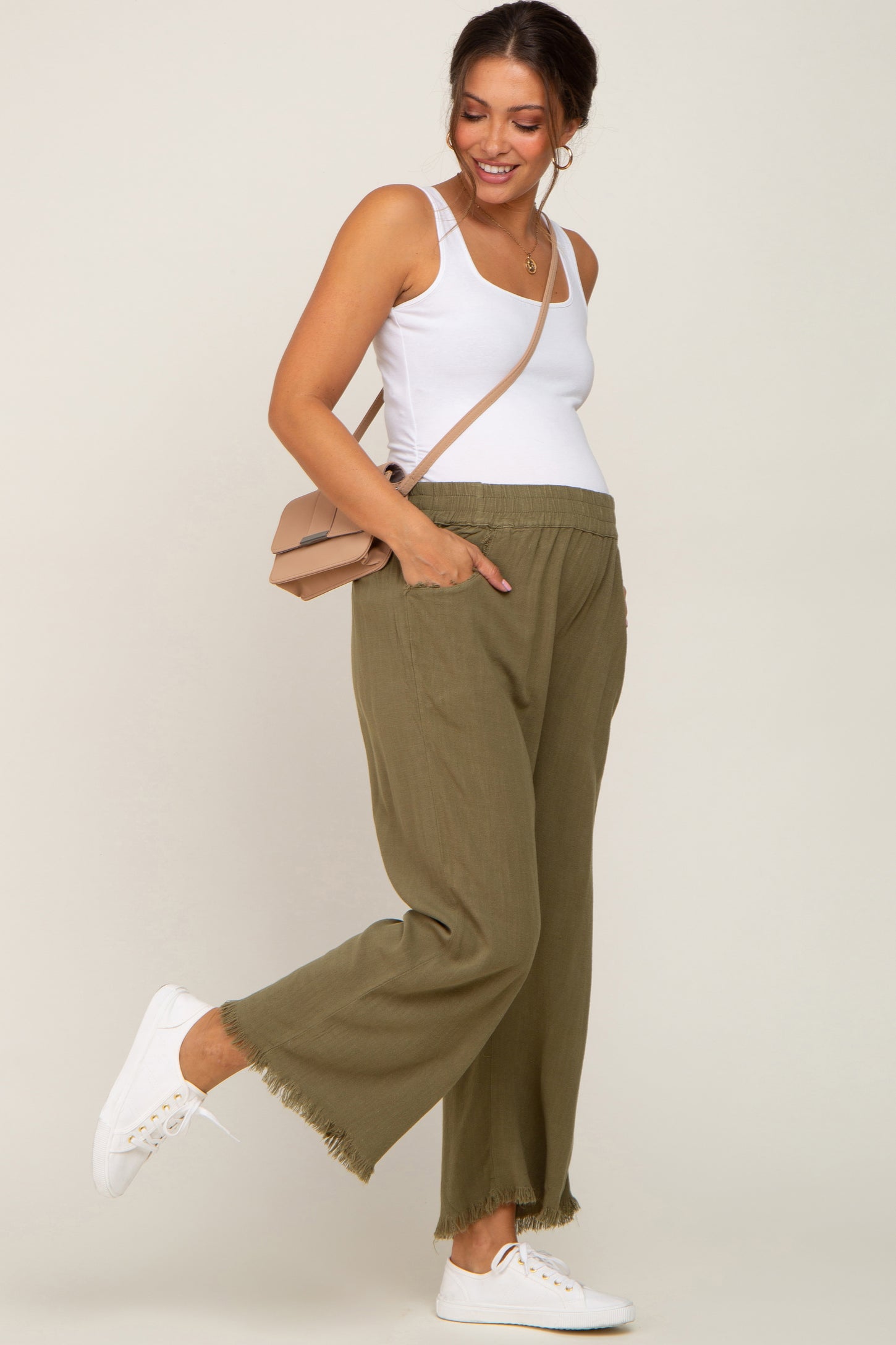 woman wearing a white tank top and olive green maternity wide-leg pants with fringe detailing, showcasing her baby bump