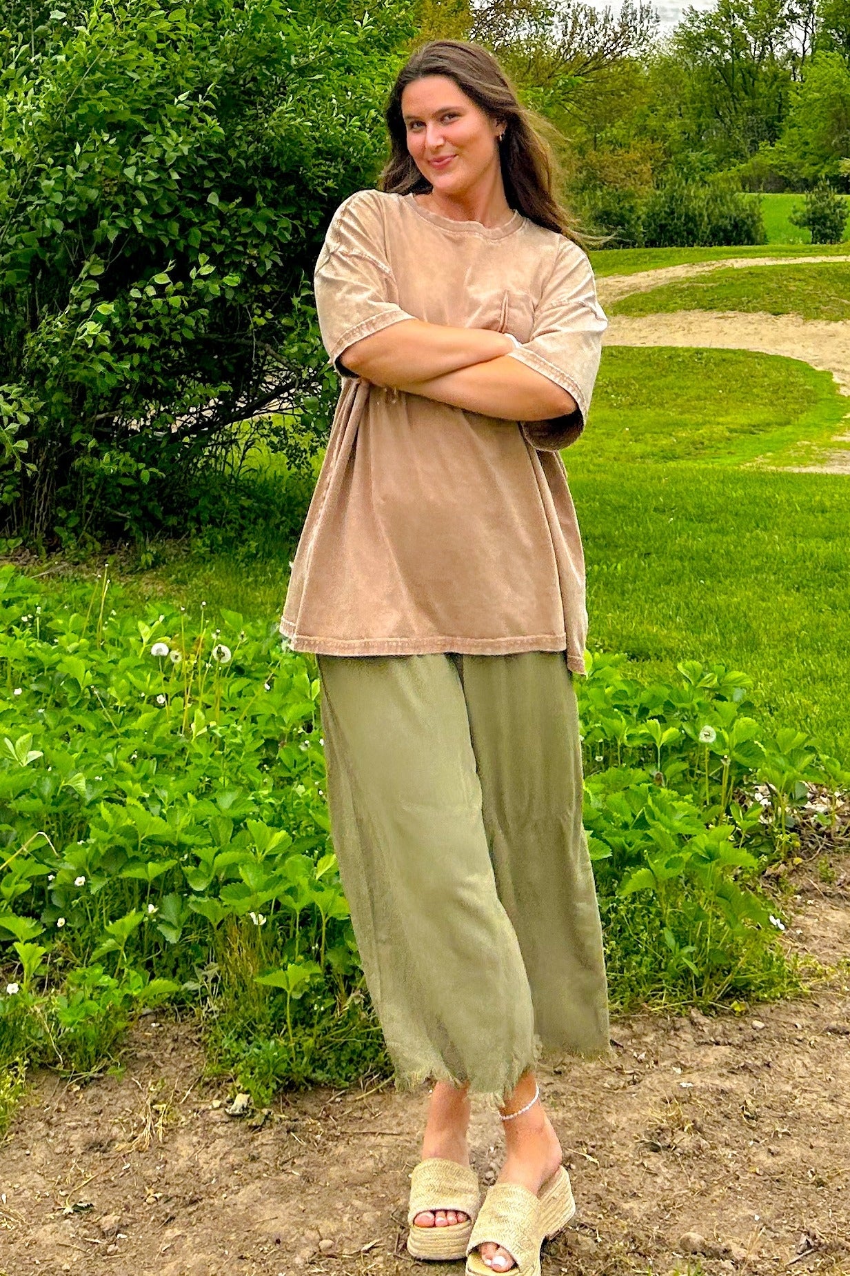 woman wearing a oversized beige t-shirt and green flowy pants standing outdoors with arms crossed