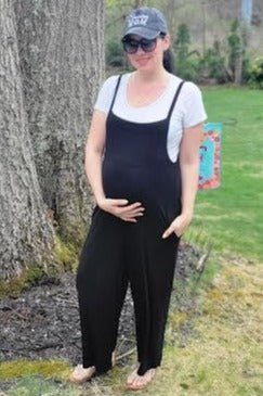 woman wearing a black maternity jumpsuit, holding her baby bump, styled with a white t-shirt and a cap