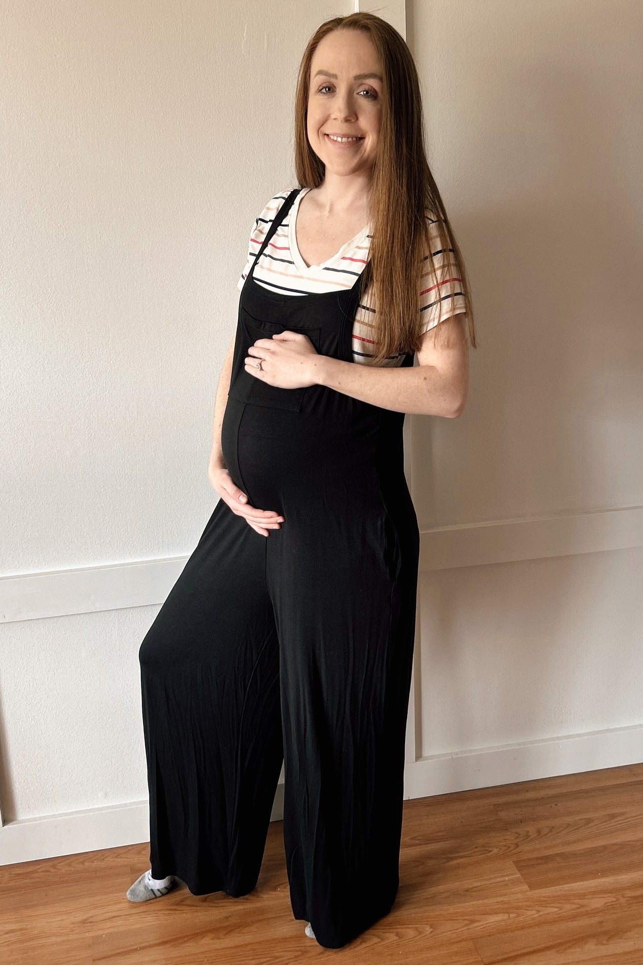 woman wearing a black maternity jumpsuit over a striped shirt, showing a baby bump while posing indoors