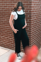 woman wearing a black maternity jumpsuit over a white t-shirt, showing her baby bump while standing against a brick wall