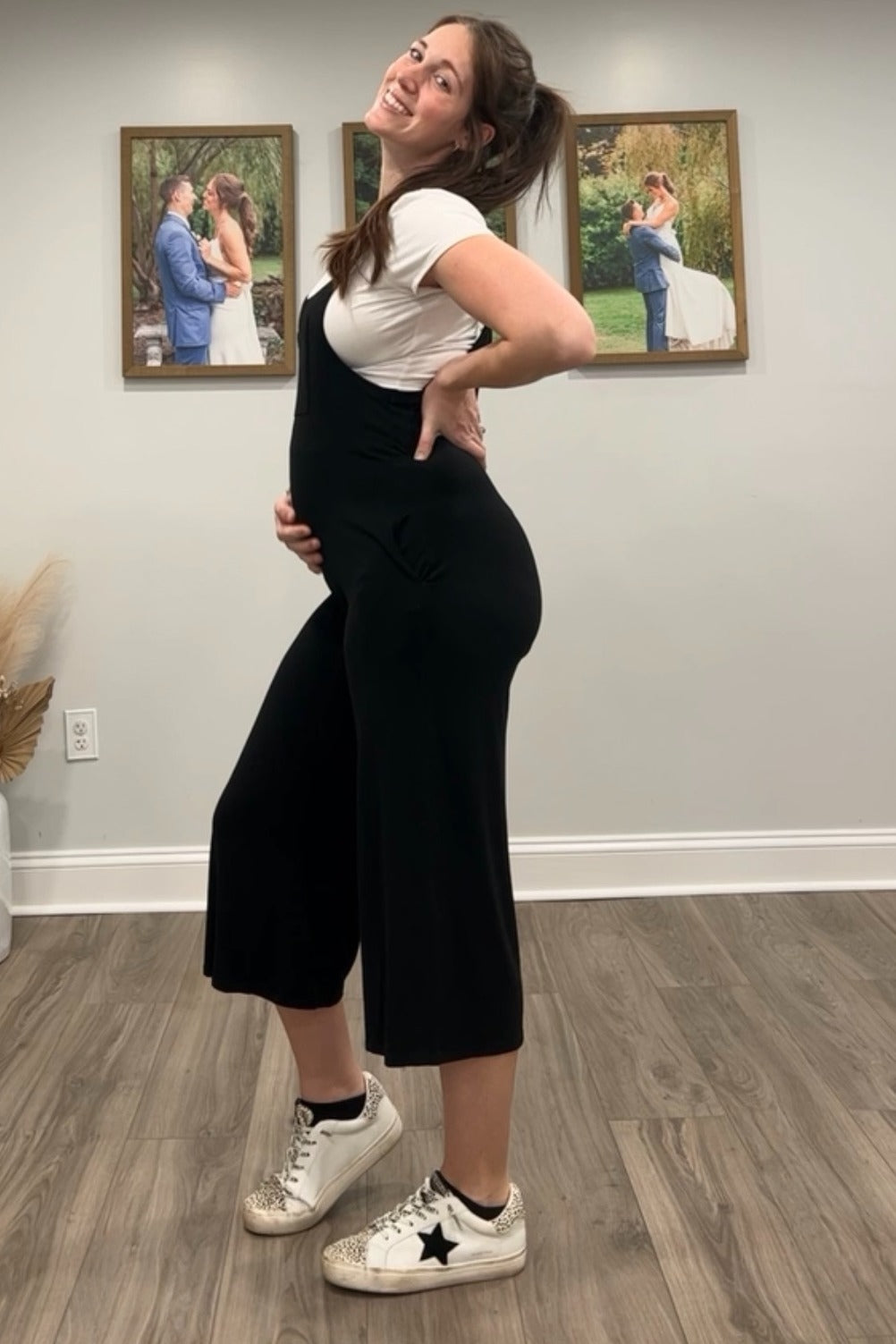 woman wearing a black maternity jumpsuit with a white t-shirt underneath and holding her baby bump