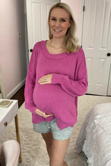 woman wearing a bright pink oversized maternity sweater and grey shorts, holding her baby bump