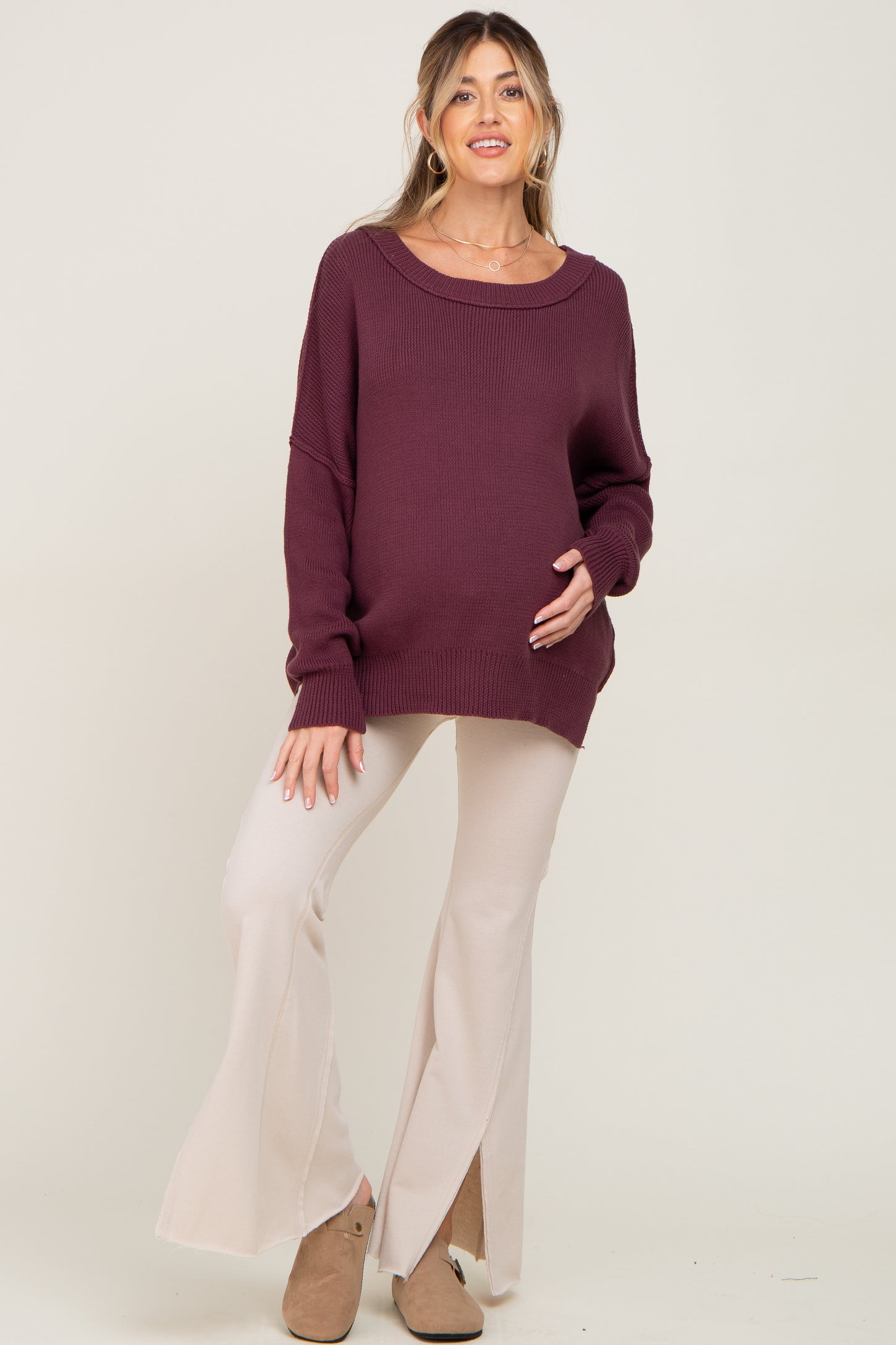 woman wearing a burgundy oversized maternity sweater and cream flared maternity pants, holding her baby bump