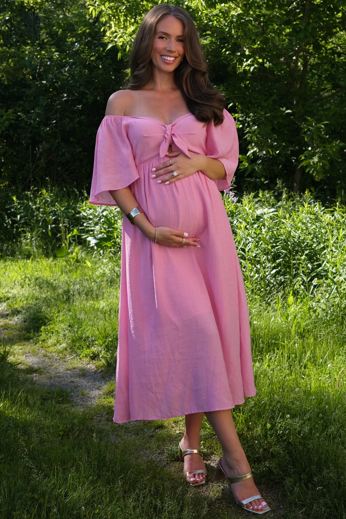 woman wearing a pink off-the-shoulder maternity dress with tie front detail and holding her baby bump in a green outdoor setting