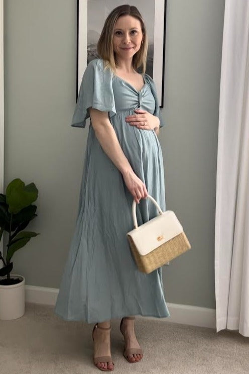 woman wearing a light blue maternity maxi dress and holding her baby bump, carrying a white handbag