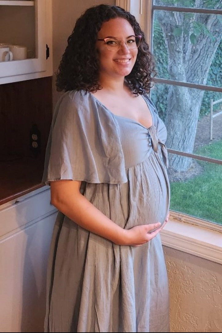 woman wearing a dusty blue maternity dress with a tie front, cradling her baby bump while standing by a window