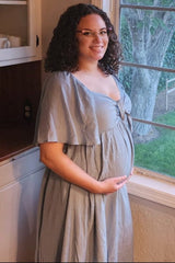 woman wearing a dusty blue maternity dress with a tie front, cradling her baby bump while standing by a window