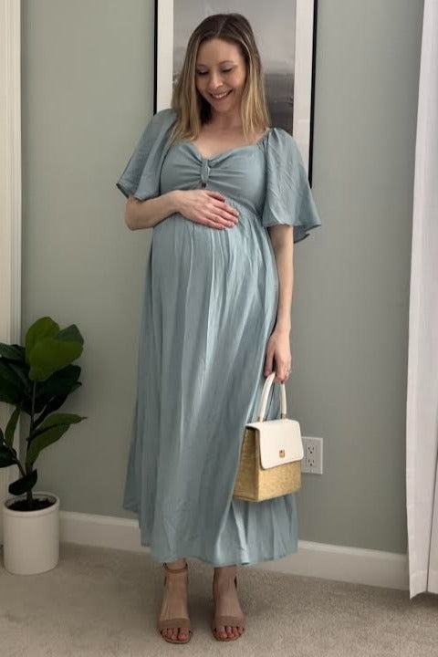 Pregnant woman in a light blue maternity maxi dress holding a straw handbag and smiling