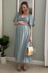 Pregnant woman in a light blue maternity maxi dress holding a straw handbag and smiling