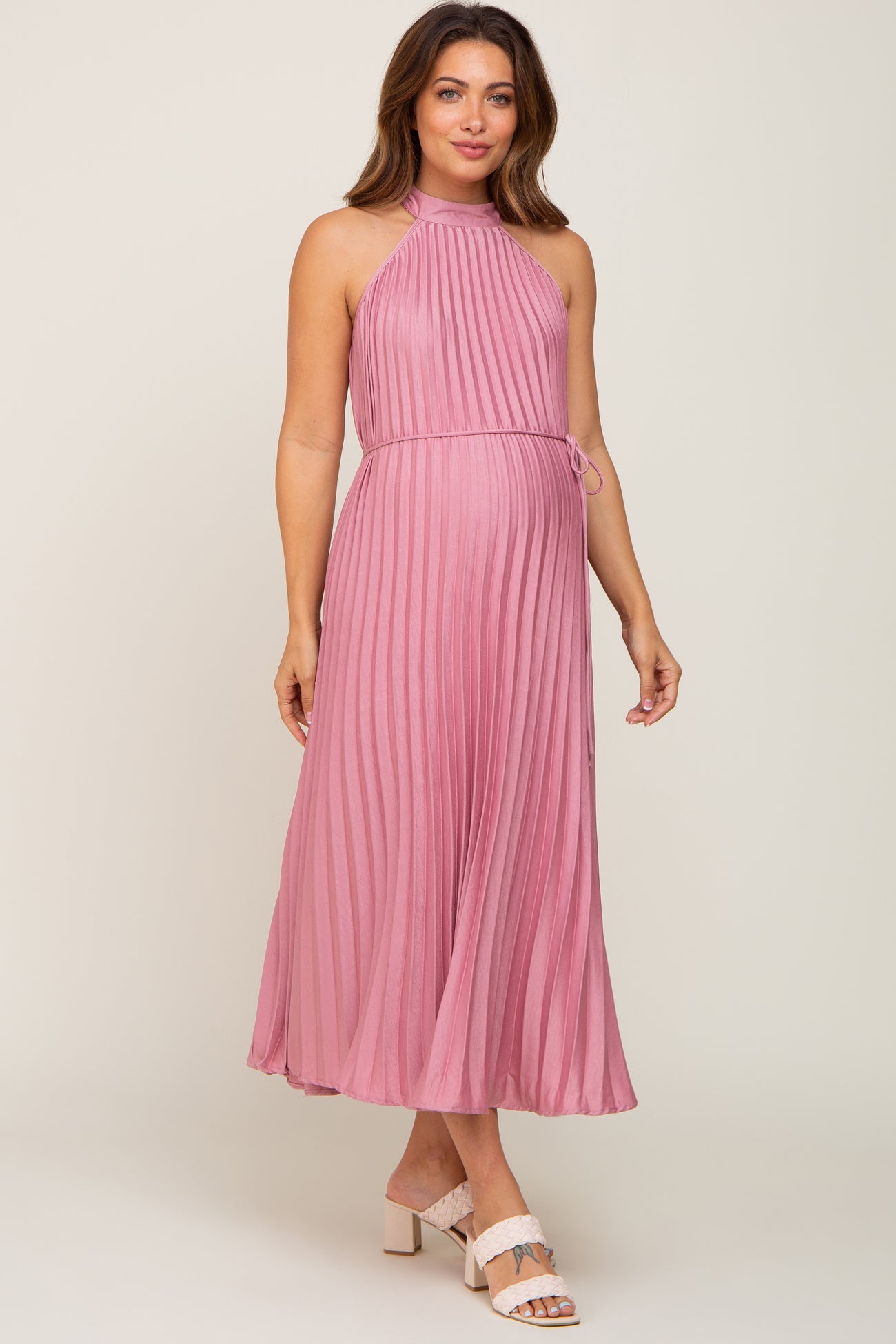 woman wearing a pink pleated maternity maxi dress with a halter neckline and a tie waist, showcasing her baby bump