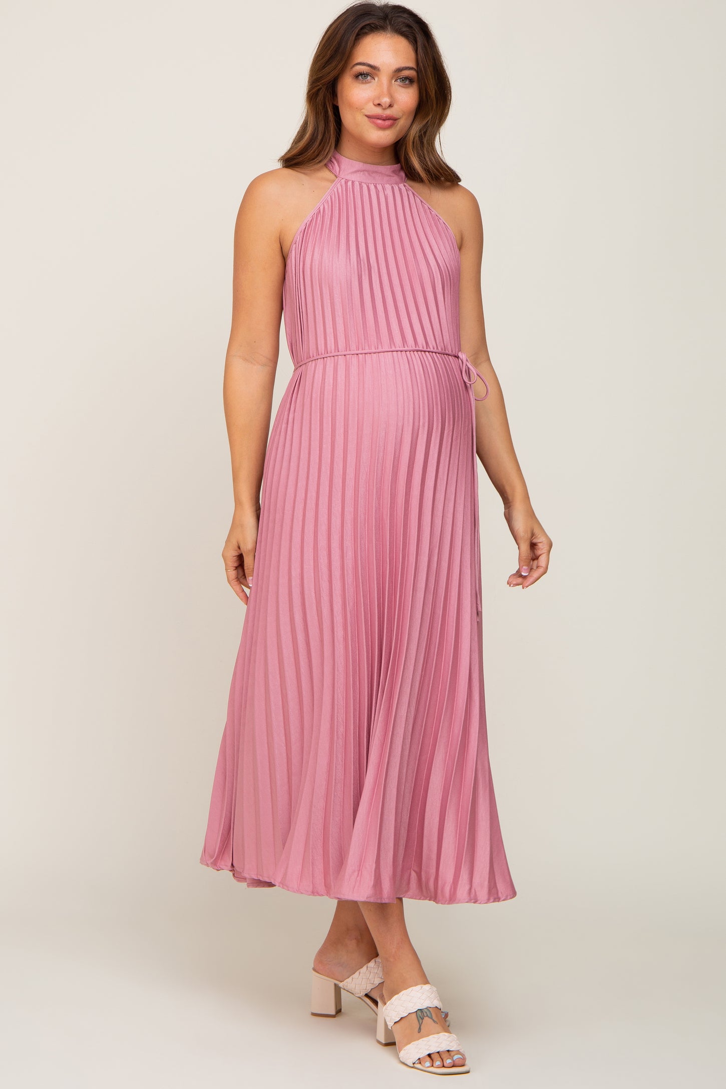 woman wearing a pink pleated maternity maxi dress with a halter neckline and a tie waist, showcasing her baby bump