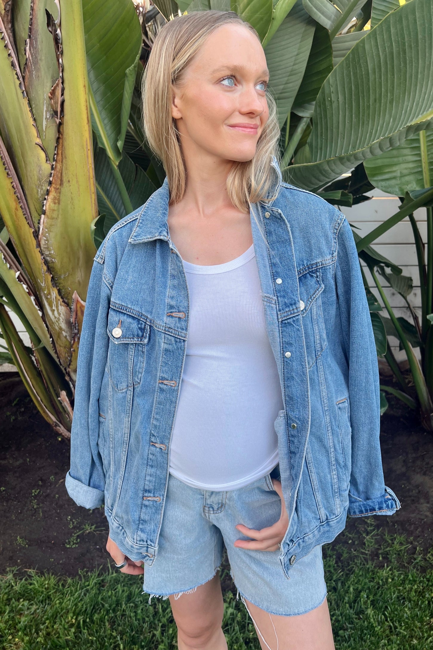 woman wearing a light blue maternity denim jacket over a white tank top, with a visible baby bump and denim shorts