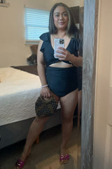 woman wearing a black one-piece swimsuit with ruffle details, holding a clutch and standing in front of a mirror