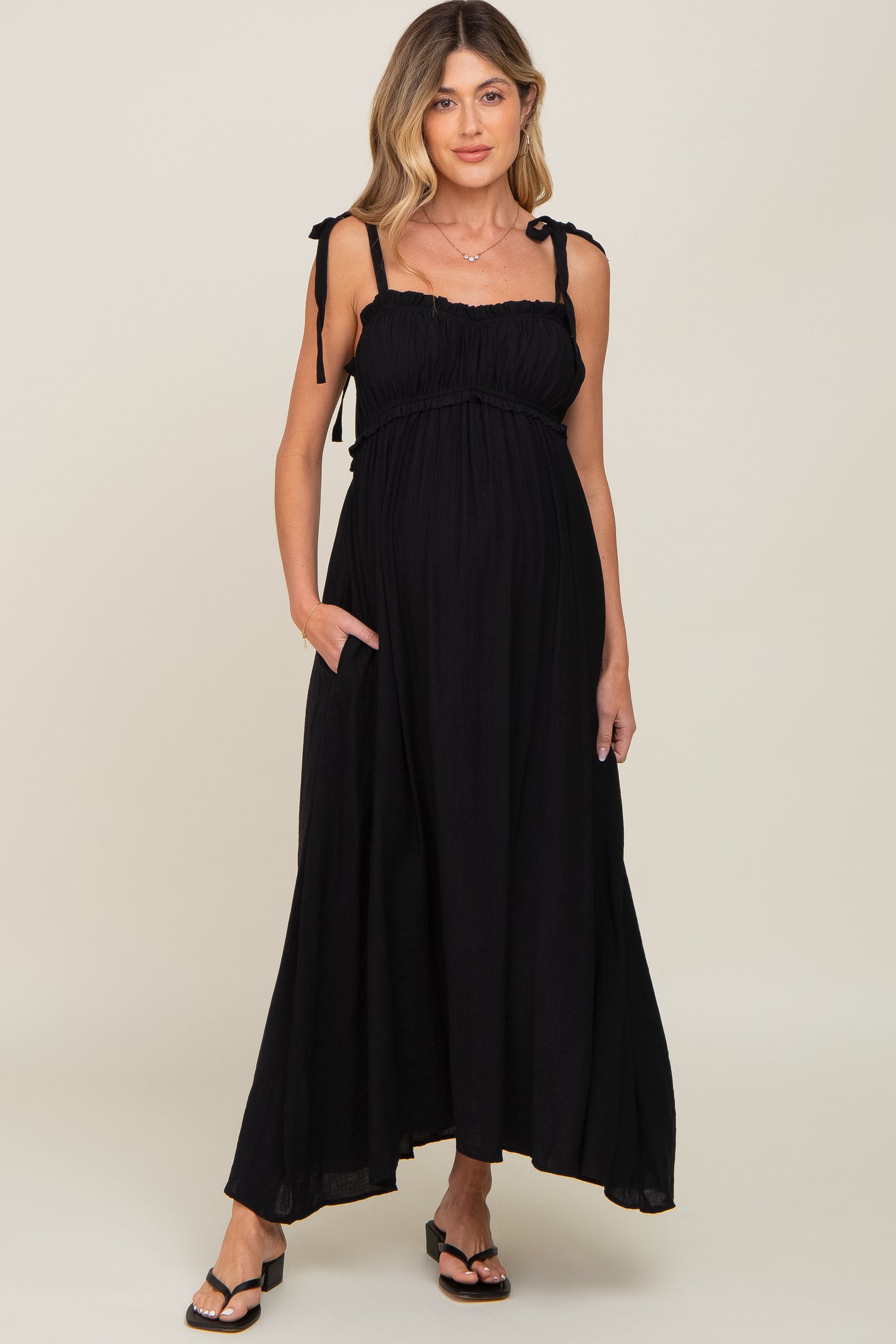 woman wearing a black maternity maxi dress with adjustable tie straps and pockets, featuring a gathered bust and flowy skirt