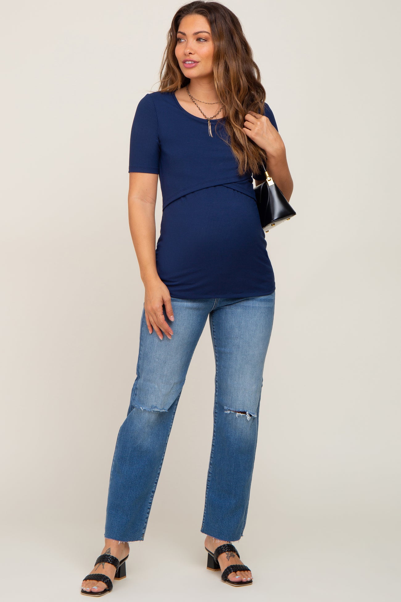 woman wearing a navy blue short sleeve maternity top with a baby bump and distressed jeans, holding a black handbag