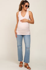 woman wearing a light pink maternity tank top and blue distressed jeans, cradling her baby bump