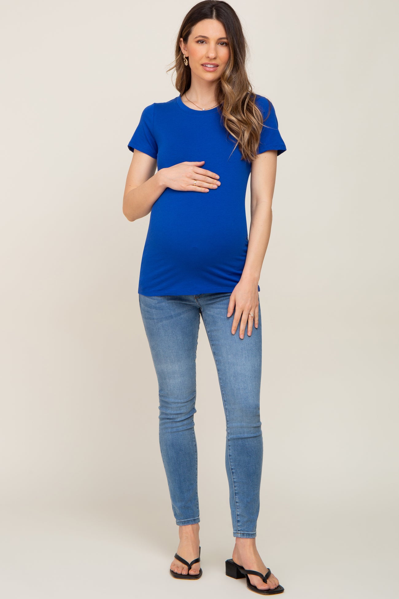 woman wearing a bright blue short sleeve maternity top and light blue skinny jeans, holding her baby bump