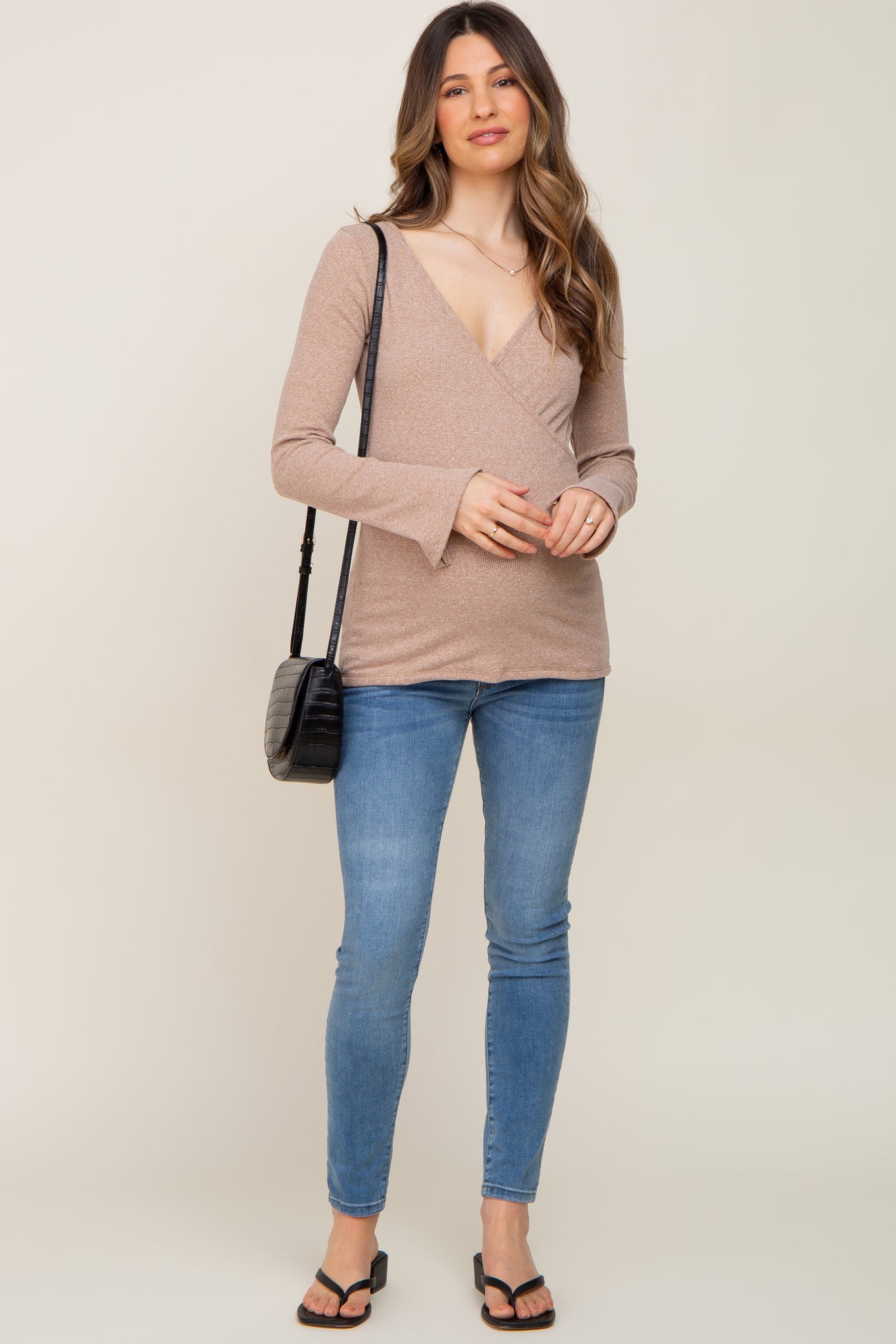 woman wearing a light brown long sleeve wrap maternity top with bell sleeves and blue skinny jeans, holding a black crossbody bag