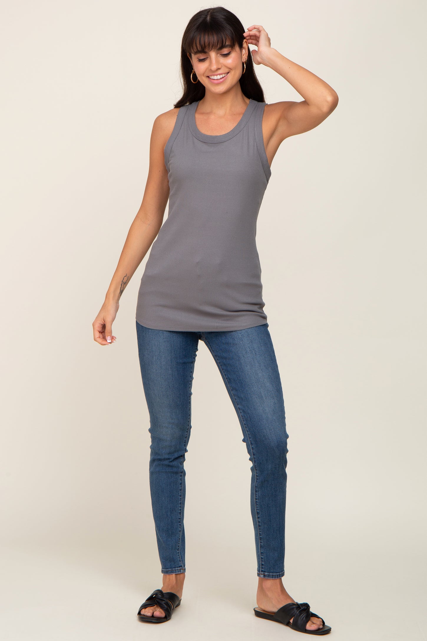 woman wearing a gray ribbed tank top and fitted blue jeans, standing with one hand on her hip