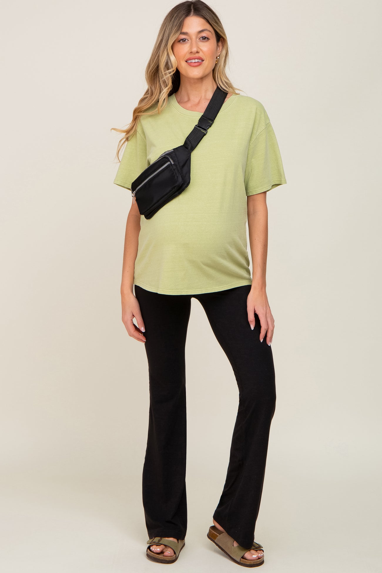 woman wearing a light green oversized maternity t-shirt and black flared pants, and posing with a crossbody bag and visible baby bump