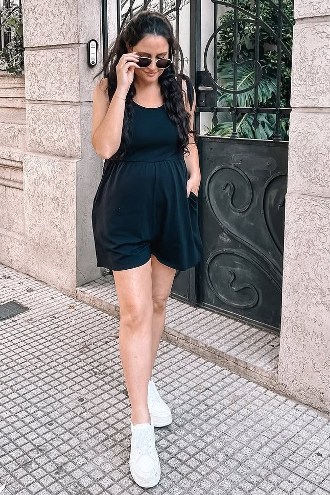 woman wearing a black maternity romper with pockets, styled with sunglasses and white sneakers, walking outdoors