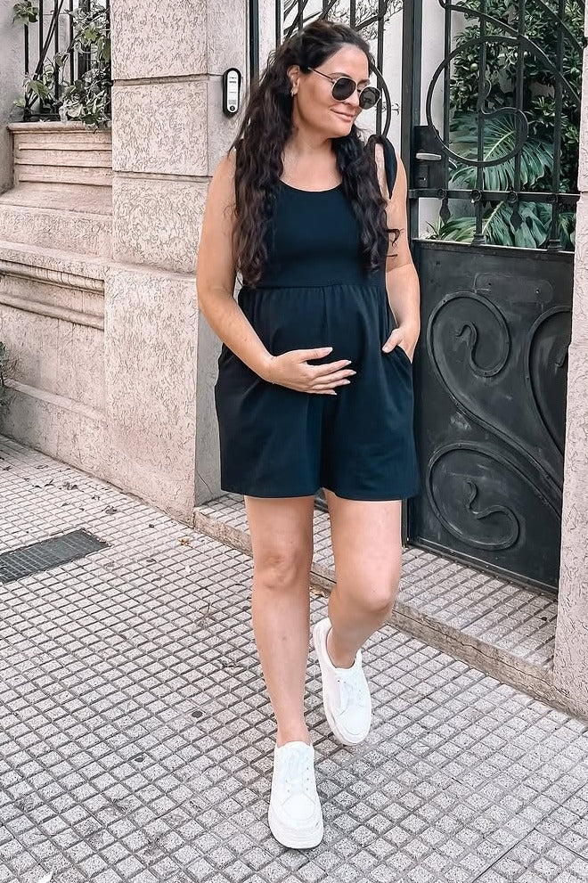 woman wearing a black maternity romper with pockets and holding her baby bump while standing outside