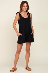 woman wearing a black sleeveless maternity romper with tie straps and pockets standing against a neutral background