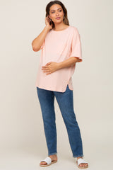 woman wearing a light pink oversized maternity top with a pocket and holding her baby bump, paired with blue jeans
