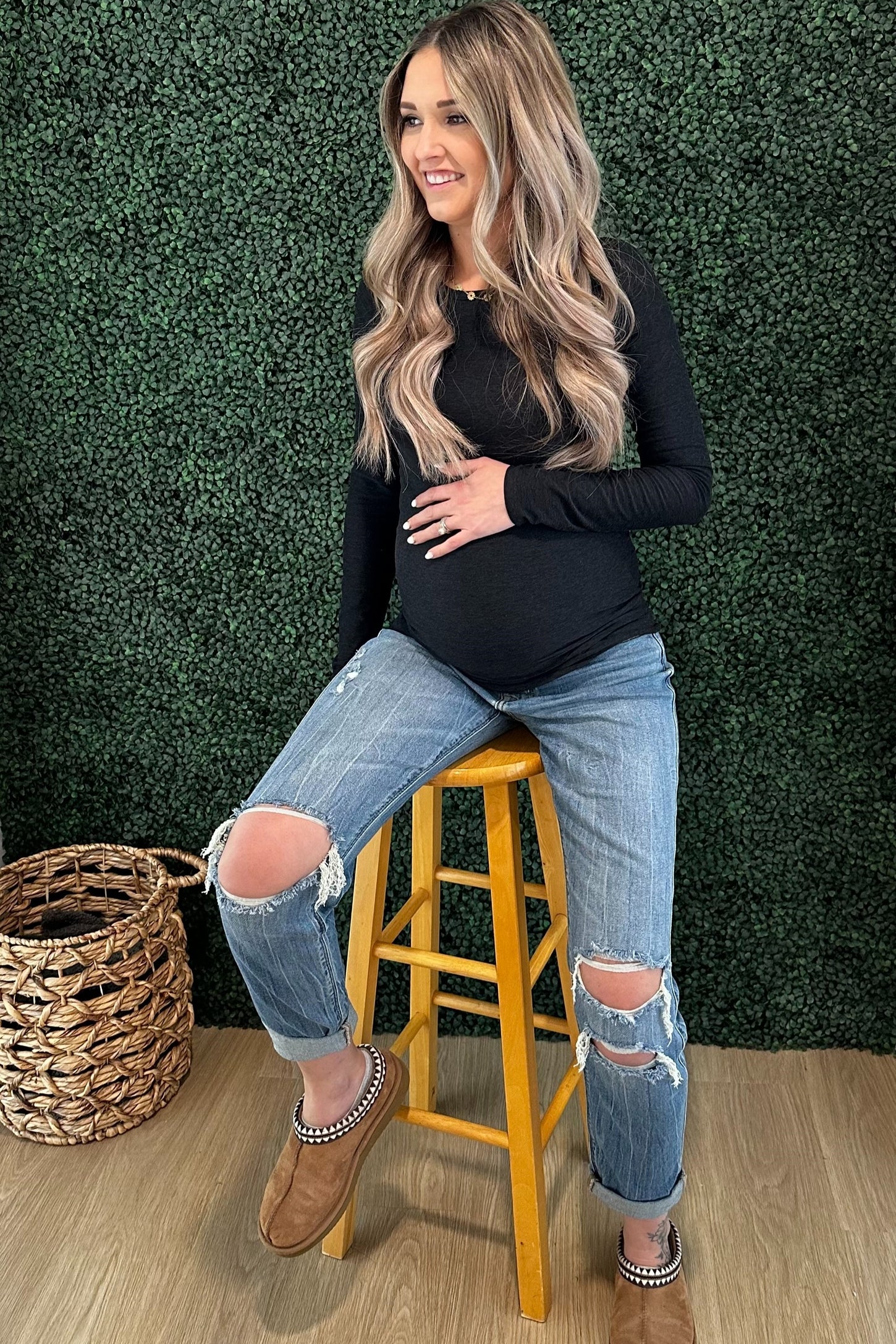 woman wearing a black long sleeve maternity top and distressed jeans, sitting on a stool with visible baby bump