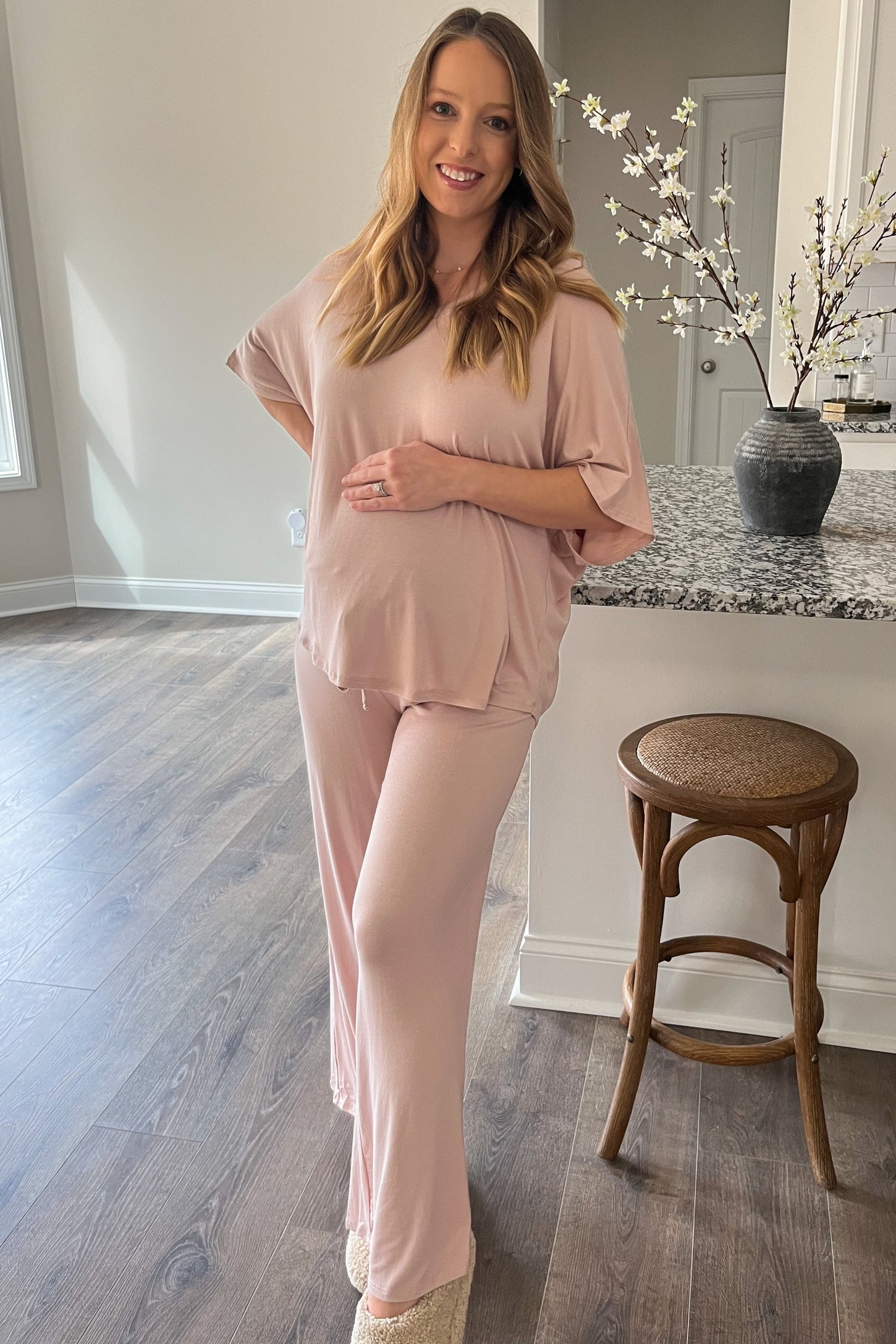 woman wearing a light pink maternity top and matching pants, holding her baby bump while standing in a well-lit room