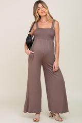 woman wearing a brown maternity jumpsuit with smocked bodice and wide legs, holding a black handbag and showcasing her baby bump