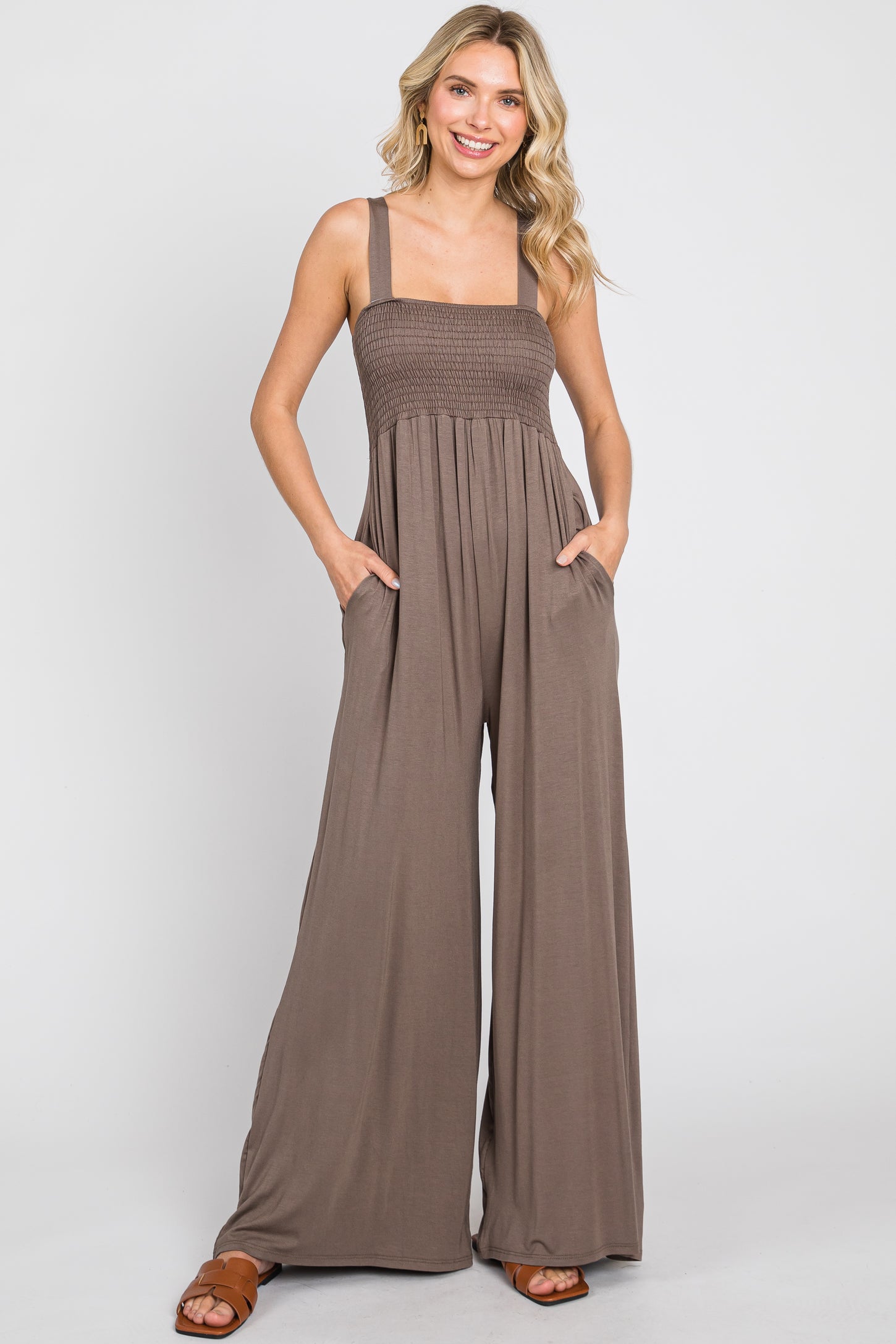 Woman wearing a brown smocked wide-leg jumpsuit with pockets and brown sandals against a white background