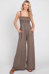 Woman wearing a brown smocked wide-leg jumpsuit with pockets and brown sandals against a white background