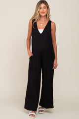 woman wearing a black sleeveless maternity jumpsuit with pockets and a white tank top underneath, standing and smiling