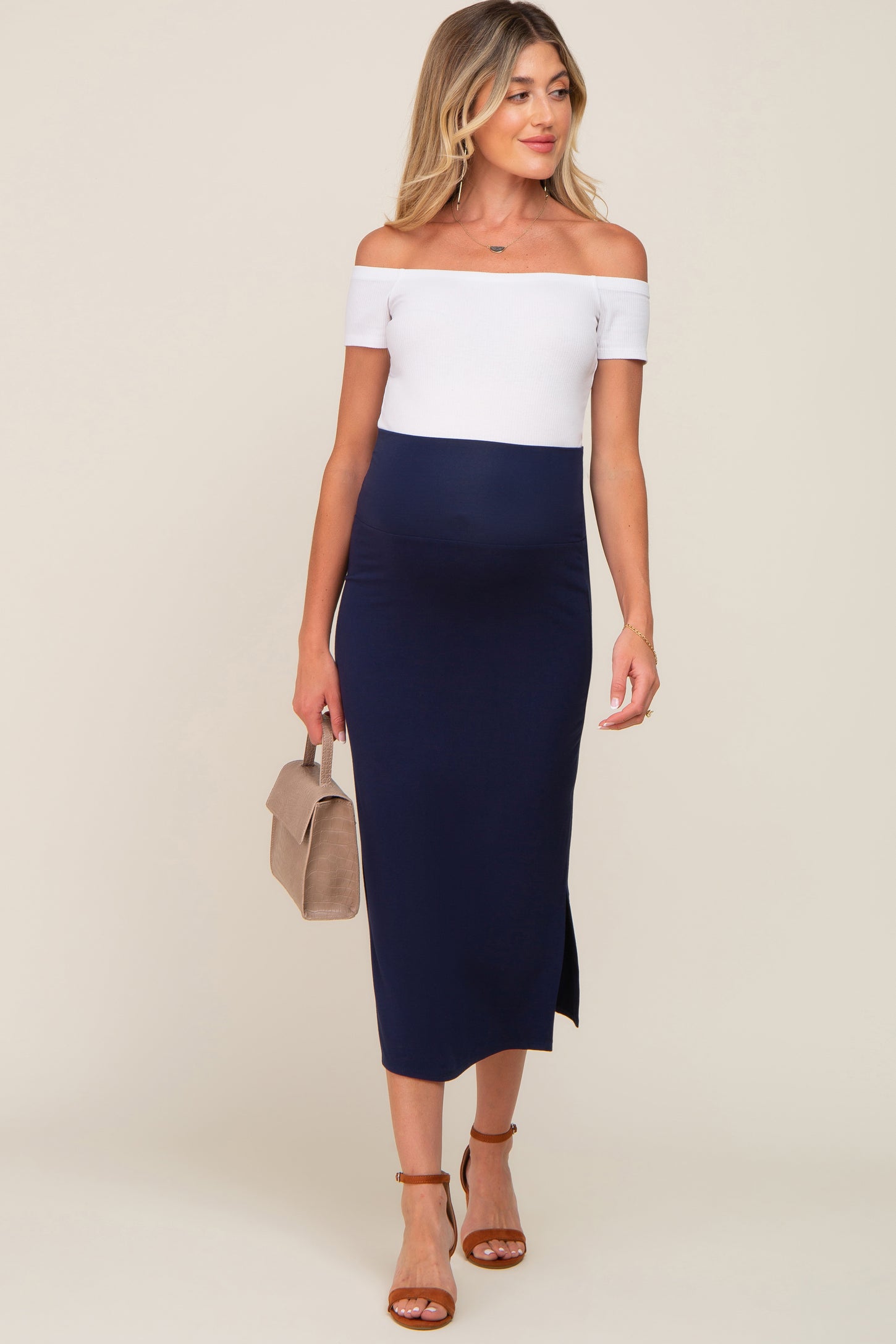 woman wearing a navy blue off-the-shoulder maternity dress with a white top and a side slit, holding a small handbag and showing her baby bump