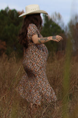 woman wearing a floral maternity dress and a wide-brimmed hat, twirling in a field with a visible baby bump