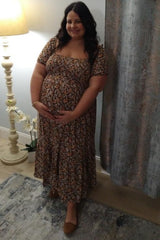 woman wearing a brown floral maternity dress and holding her baby bump while standing by a lamp