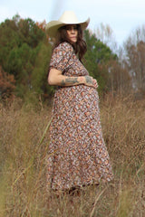 woman wearing a brown floral maternity dress with a ruffled hem and a cowboy hat, showing her baby bump in a field