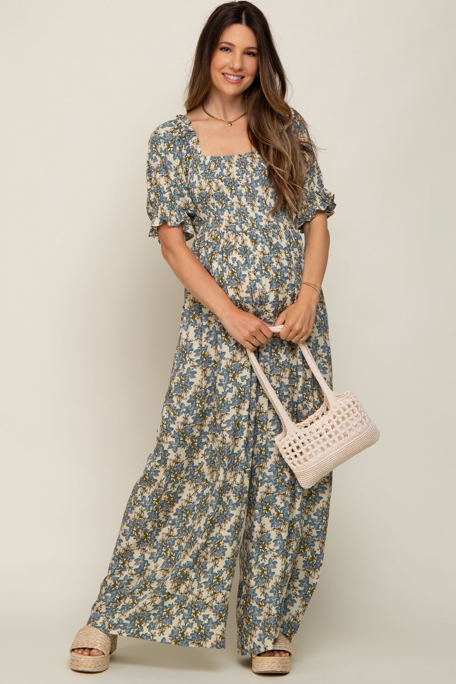 woman wearing a floral maternity maxi dress with a square neckline and puff sleeves, holding a woven handbag