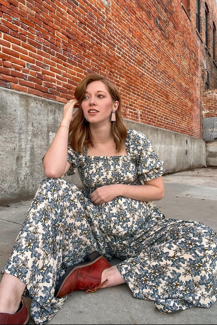 woman wearing a floral print maternity jumpsuit and showing her baby bump, seated against a brick wall