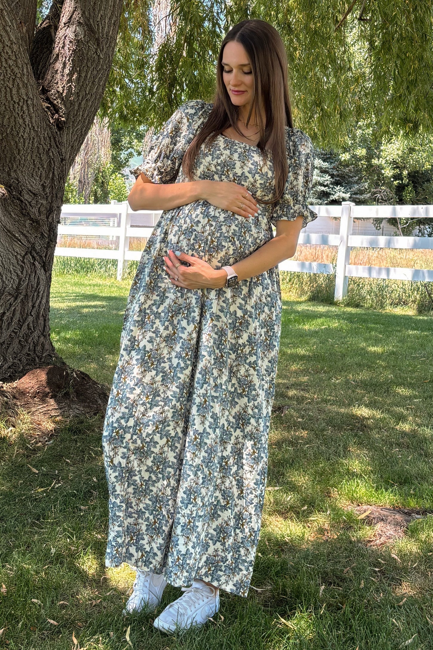 woman wearing a floral maxi maternity dress and holding her baby bump while standing in a grassy area under a tree