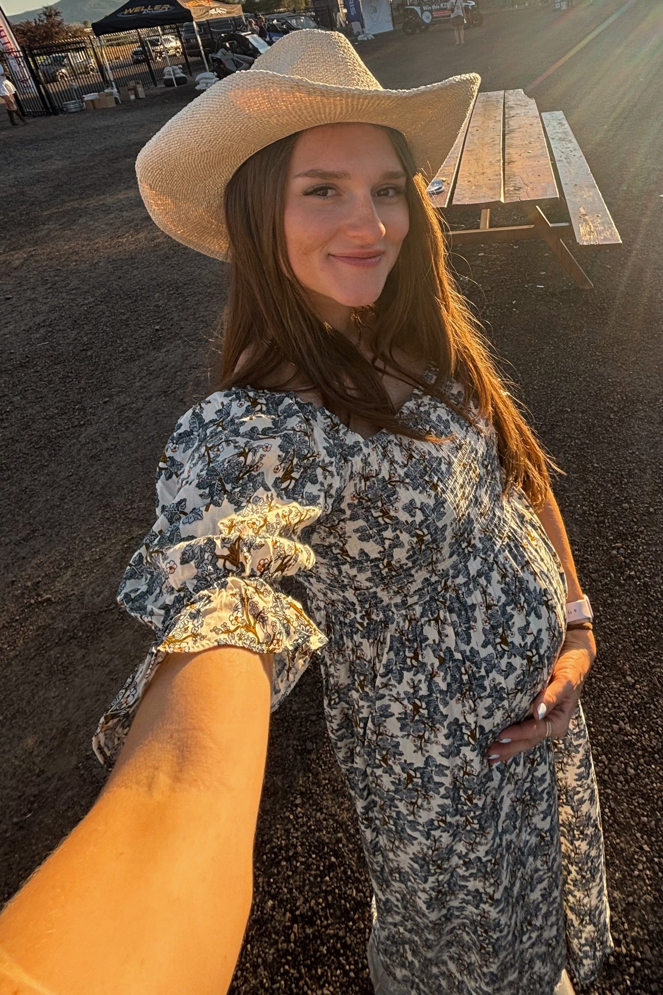 pregnant woman in floral dress and straw cowboy hat taking a selfie outdoors at sunset