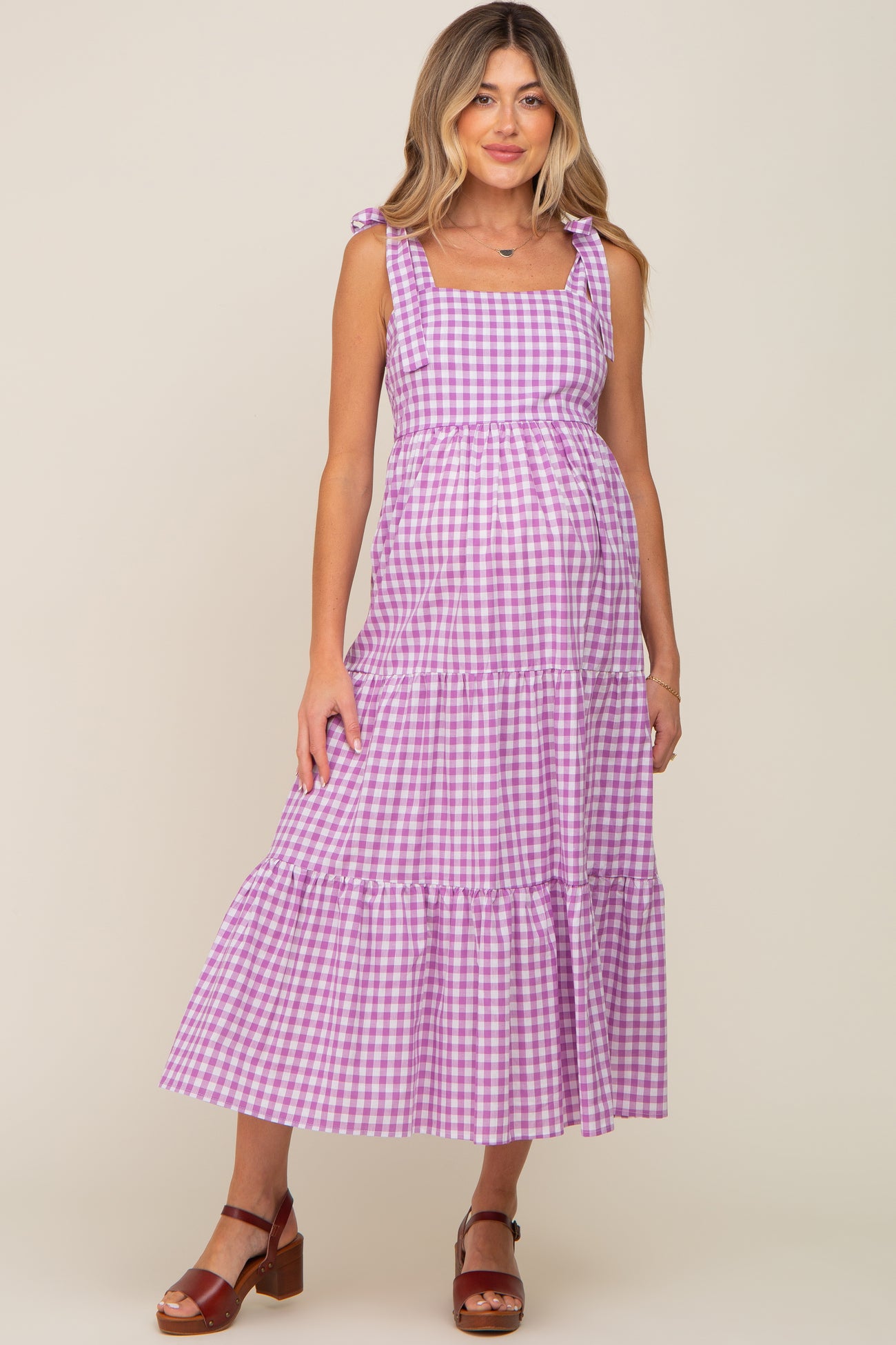 woman wearing a pink and white checkered maternity dress with a tiered skirt and tie straps, showing her baby bump