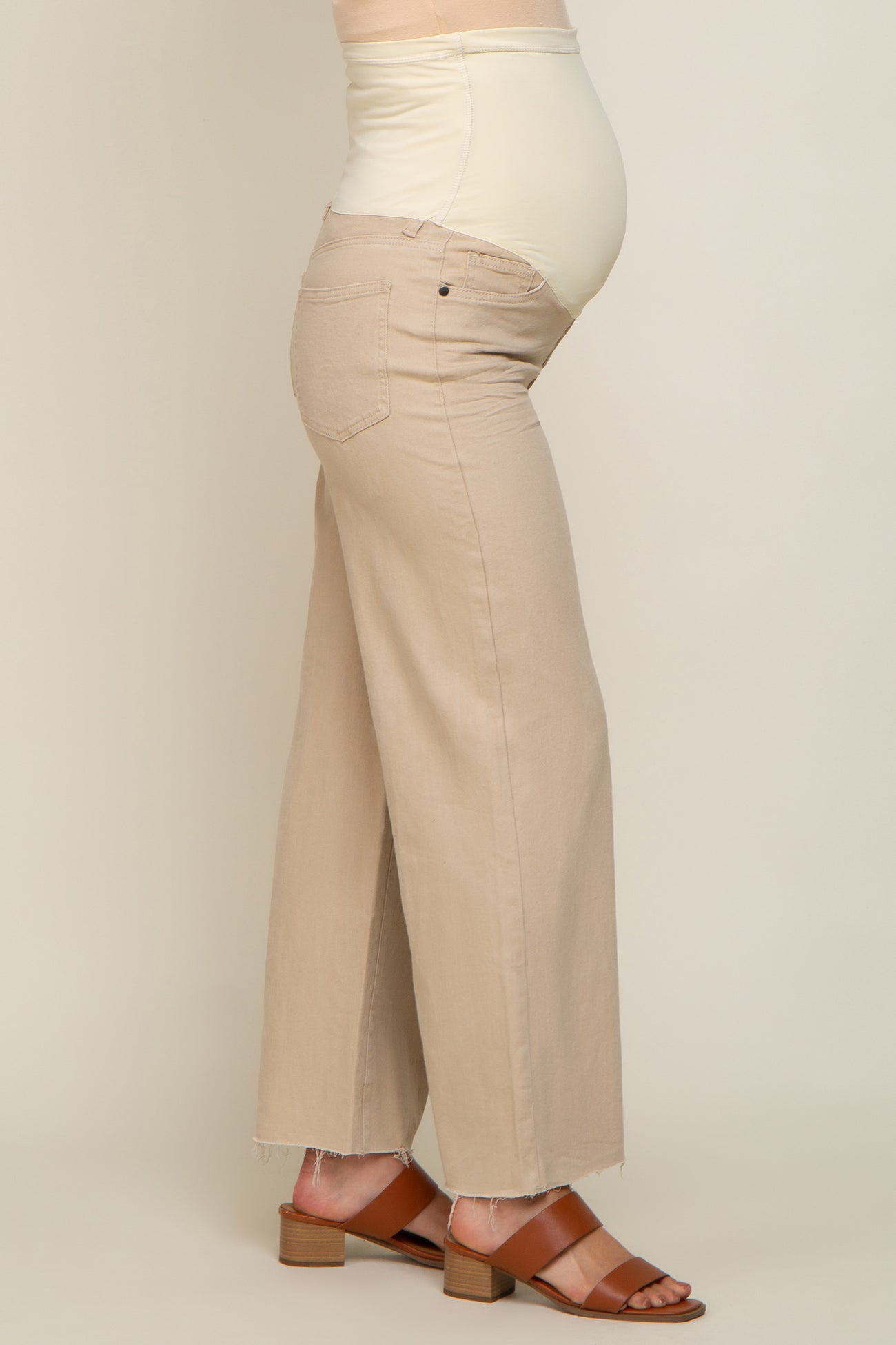 woman wearing a beige maternity pant with a stretchy belly panel, showing her baby bump, paired with brown block-heeled sandals