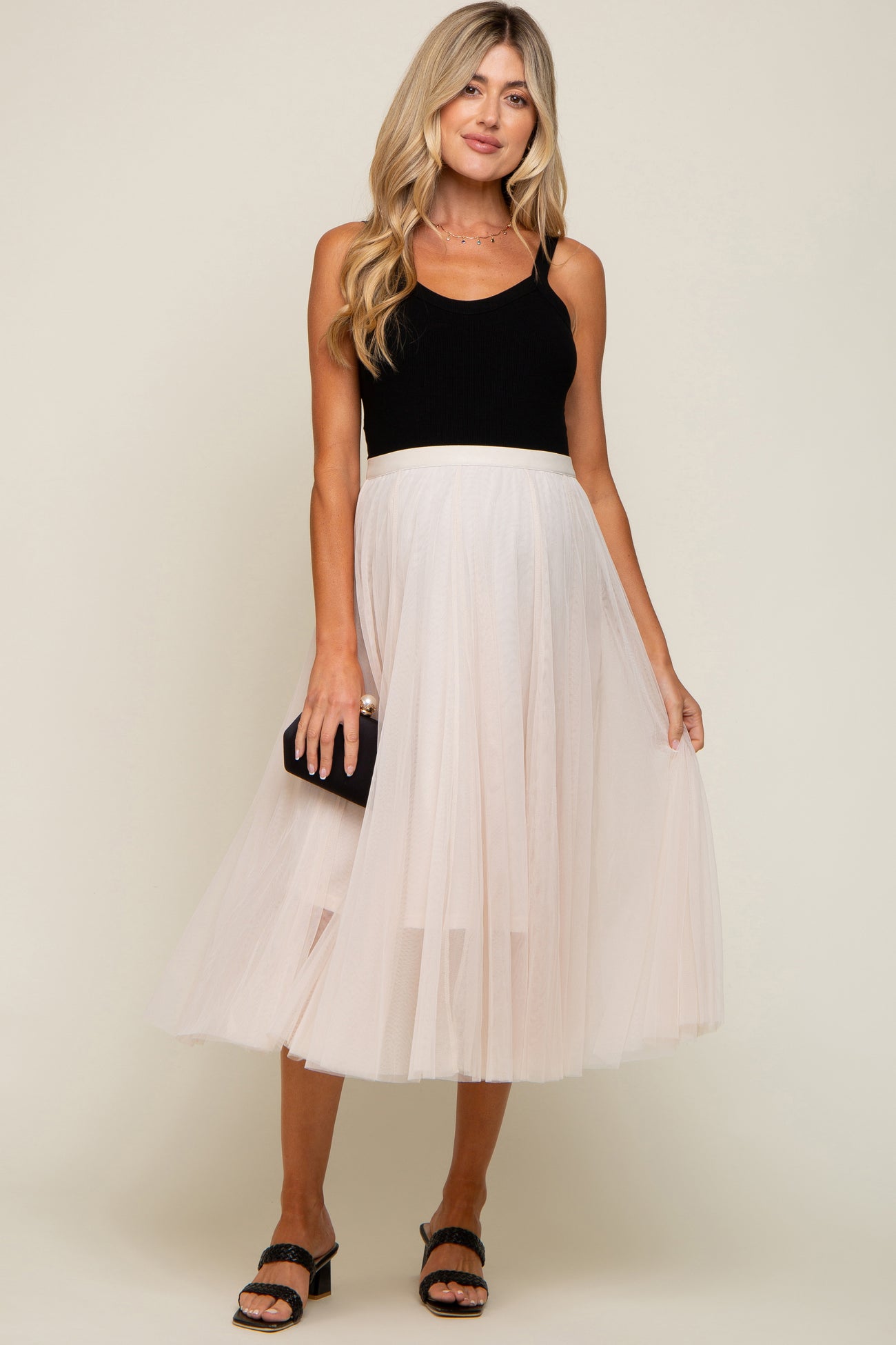 woman wearing a black ribbed maternity tank top and a light pink tulle midi skirt, holding a clutch bag