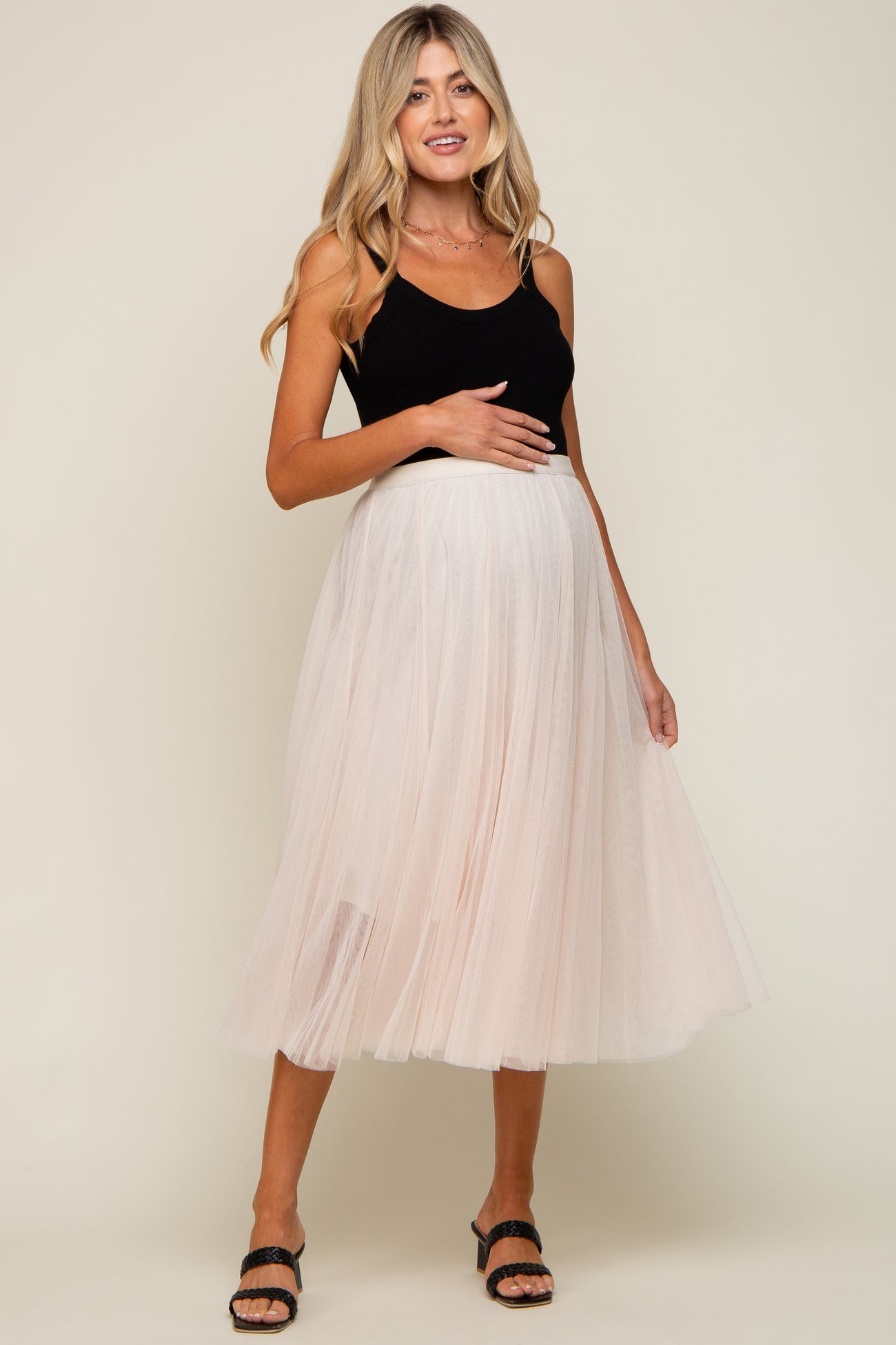 woman wearing a black tank top and a light pink tulle maternity skirt, gently holding her baby bump